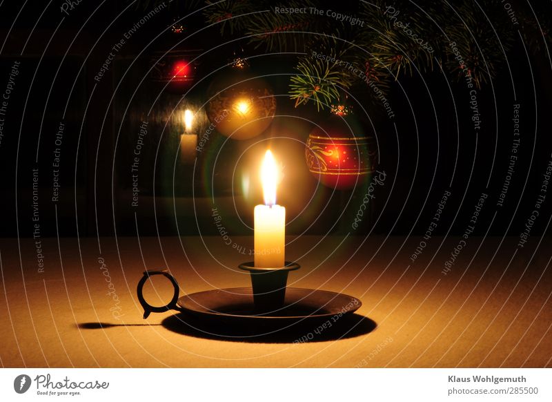 A lone candle glows, in the background fir branches on which Christmas tree baubles shine in the candlelight. The candle flame creates lens flares in the lens.