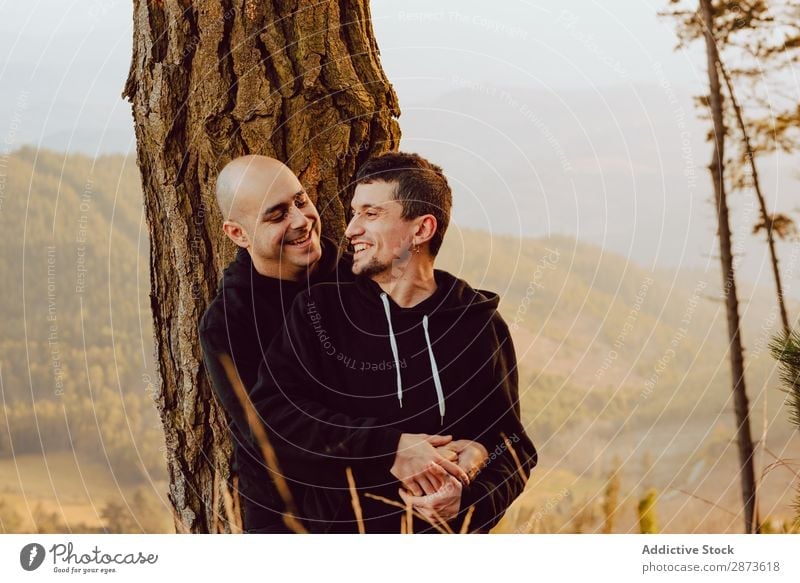 Smiling men hugging near tree in forest on hill Homosexual Couple Embrace Tree Hill Forest Love embracing Valley Picturesque Vantage point romantic Mountain