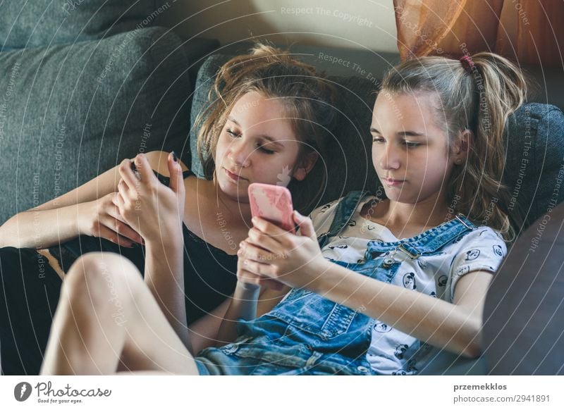 Young women using mobile phones watching music clip, texting, messaging. Teenagers using the smartphones, sitting on sofa at home. Using technology devices. Girls wearing summer clothing