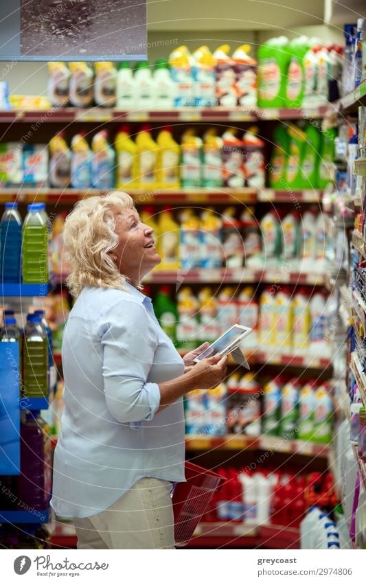 A smiling middle aged woman in a light blue shirt is standing in a household section of a supermarket. She is holding a tablet and a red shopping basket in her hands. A woman is looking at the shelves, searching for something particular