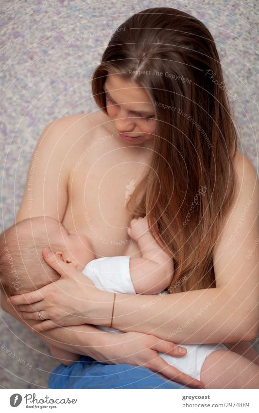 A long haired young woman is breastfeeding a baby boy in a white onesie, holding him in arms. She is looking at son with a gentle smile Nutrition Child