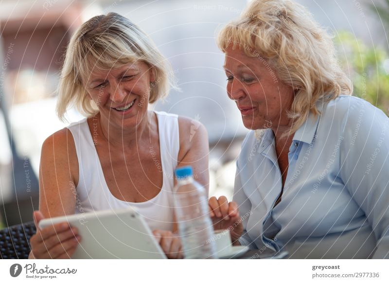 Two middle-aged women sitting in a street cafe and smiling to something in their pad Drinking water Lemonade Bottle Joy Computer games To talk Human being