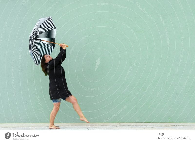 Woman with black coat and short jeans is dancing barefoot with an umbrella in her hands in front of a light green wall Human being Feminine Adults 1