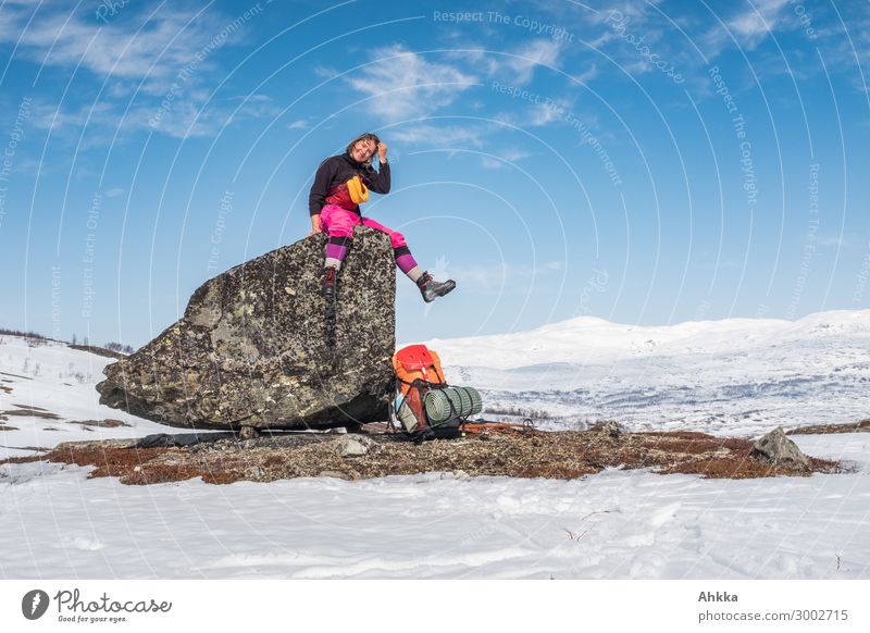 Young woman with colorful pants sits on a large stone lying on a snow-free surface and looks childlike unselfconsciously into the camera, orange backpack, cheerful, free