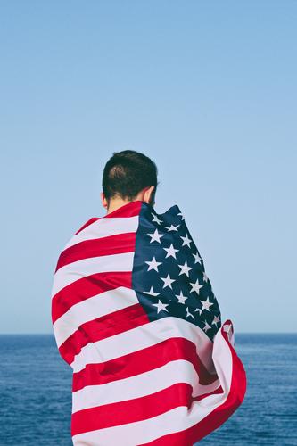 Man from behind wrapped in American Flag against blue sky Happy Freedom Ocean Feasts & Celebrations Adults Hand Sky Monument Stripe Blue Honor Independence