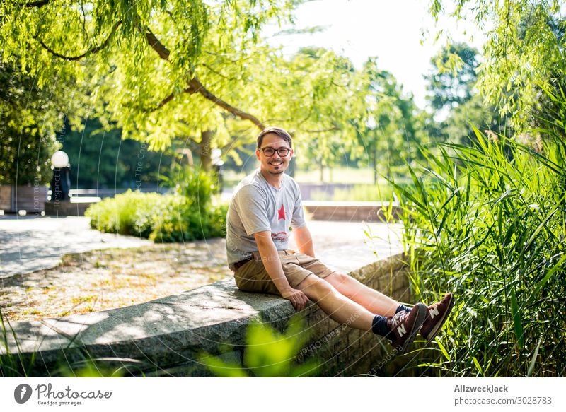 young man sits on a wall and rejoices Beautiful weather Cloudless sky Blue sky Green Nature Tree Common Reed To enjoy Relaxation Break Restful Park Berlin