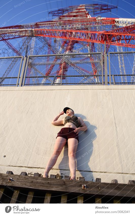 Young woman stands in bellyless top and hotpants under red and white overhead line mast on a wooden scaffold and looks into the distance Lifestyle Joy