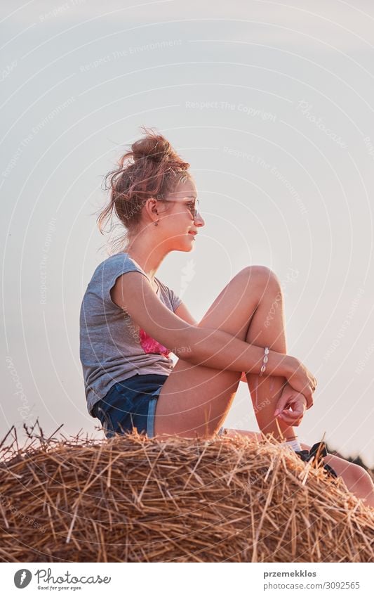 Teenage girl sitting on a hay bale at sunset relaxing while spending summertime in the village. Candid people, real moments, authentic situations Lifestyle Joy