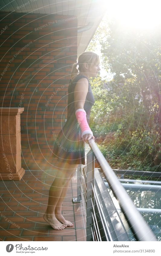 Portrait of a young slender woman with forearm in pink plaster standing barefoot on a balcony railing looking into the distance Style Joy pretty Well-being