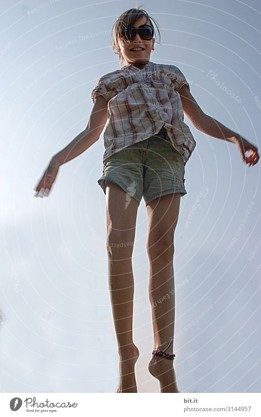 Girl with sunglasses, short jeans and long legs, jumps happily in summer, happily up on the trampoline, with blue sky in the background. Leisure and hobbies