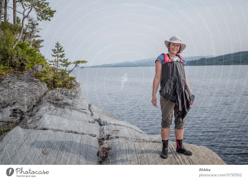 Young woman with hat stands on the edge of a rock on the shore of a lake and looks at the camera Adventure vacation Lake Rock Kayaker Nature bank Water