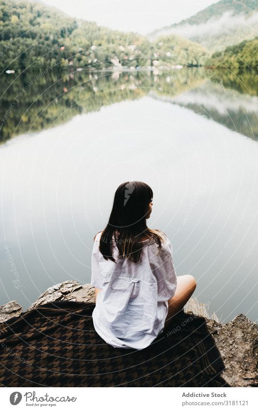 Woman sitting on blanket near lake and mountains Sit Blanket Picnic Lake Mountain Picturesque Water Coast Surface Amazing Vantage point Hill Stone Sky Clouds