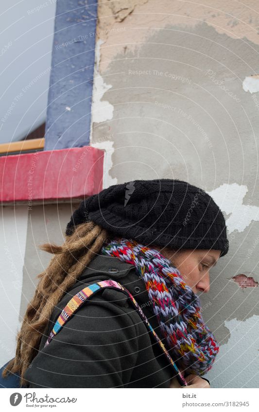 Side view in half profile of a young woman with dreadlocks, cap and colourful scarf, who is tired, exhausted, sadly leaning against an old grey house wall with a colourful window during an excursion in the city to have a quiet break.