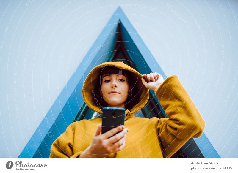 Woman with smartphone against triangle window woman using adjust hood building wall gray young female casual jacket warm exterior coat geometric shape lady