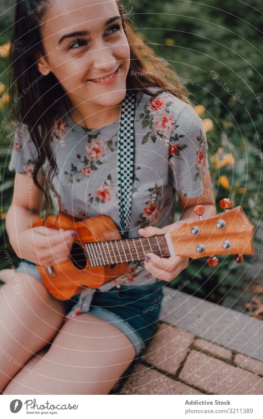 Happy modern woman playing ukulele city smiling casual sit pavement flowerbed trendy street long hair t-shirt satisfied instrument young fun leisure carefree