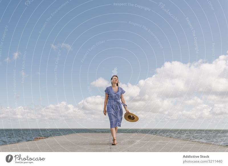 Young woman on sea wharf pier run freedom escape motion horizon straw hat landscape seaside waterfront holding hat destination bay sundress concrete quay clouds