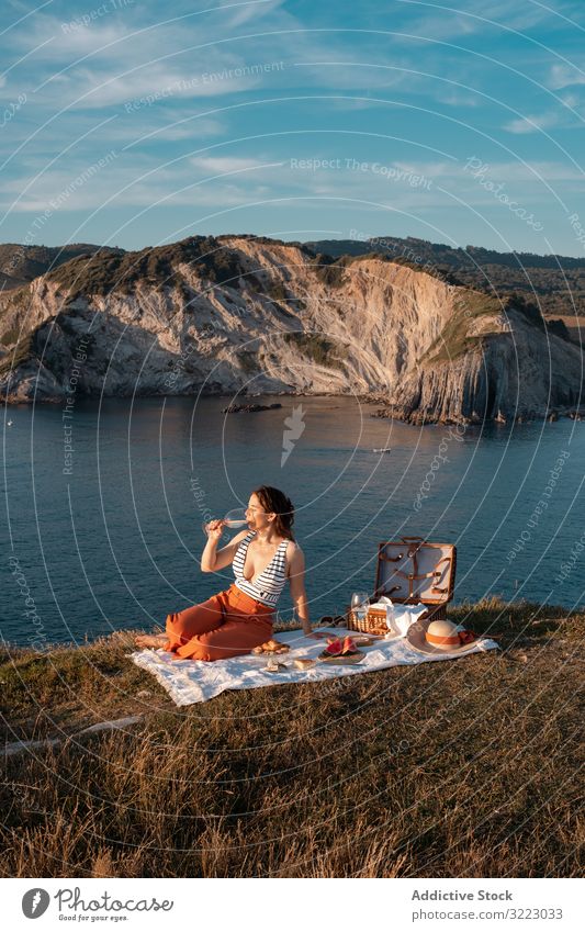 Woman with glass of drink on picnic mat looking at sea and mountains woman seaside beach summer reading leisure sky relax vacation summertime vintage fashion