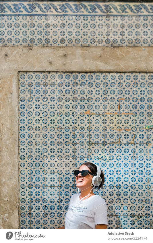 Cheerful casual woman in sunglasses standing on city street headphones stylish cheerful trendy millennial beautiful gadget music earphones device happy building