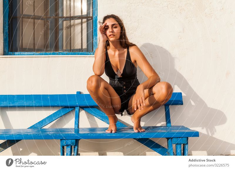 Young female relaxing on blue bench woman squat rest pensive recreation stylish casual pretty brunette slim trendy sad upset black shorts top barefoot tattooed