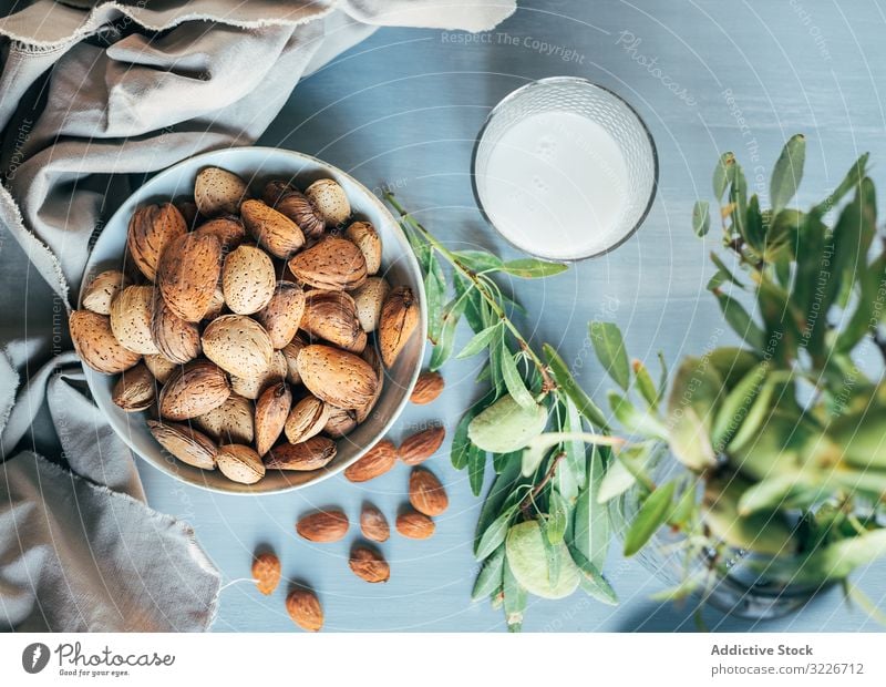 Glass of almond milk next to plate of almonds in shells on kitchen table glass nuts bowl cloth twig green plant rustic snack breakfast ingredient vegetarian