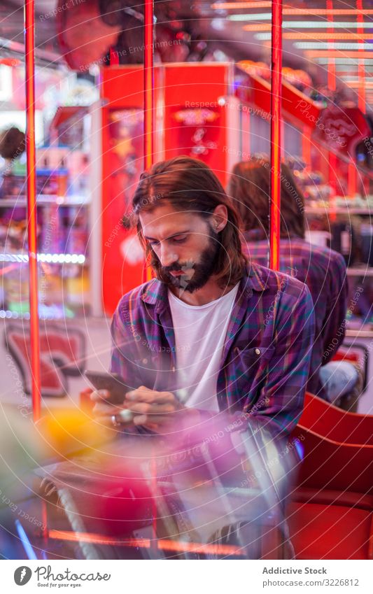 Focused hipster man texting on smartphone while resting at fairground carnival focused attentive bearded distracted alone solitude casual serious using sit