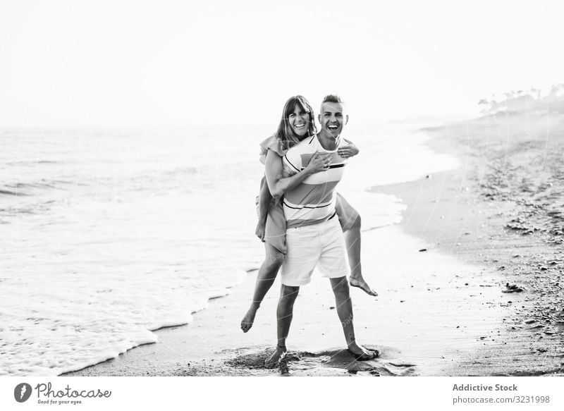 Man carrying woman on back near sea couple beach resort love piggyback ride smile happy vacation fun wave ocean water adult honeymoon summer shore coast