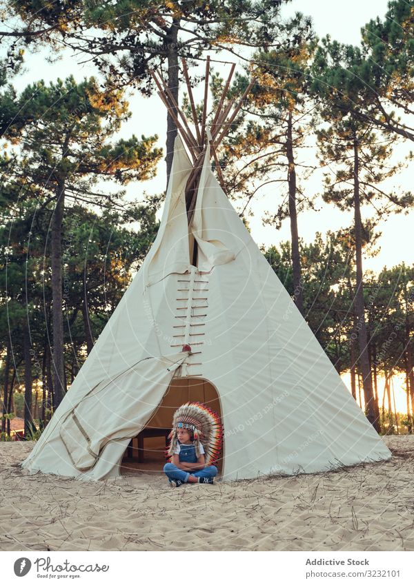 Content kid in authentic hat by tent boy wigwam indian feather content delighted overalls denim standing crossed arms portugal park leisure playhouse childhood
