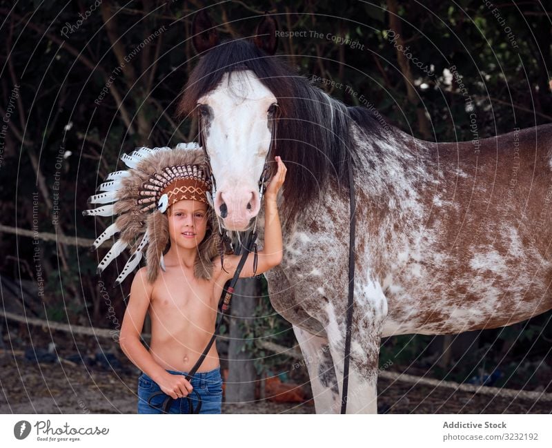 Kid in authentic headdress hugging stallion on farm boy horse caress war bonnet kind child indian costume concentrated stroke shirtless native art head wear