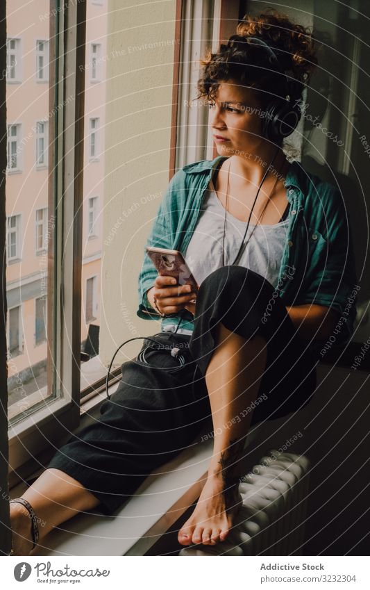Relaxed woman in headphones using smartphone at home smile pensive listen happy music browsing window sill curly attractive peaceful relaxed apartment young