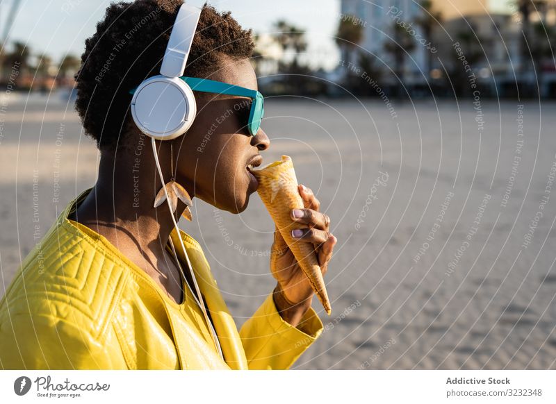 Cool female eating ice cream on beach woman trendy cool desert african american headphones jacket yellow bright sunglasses enjoy sweet food summer fun stick