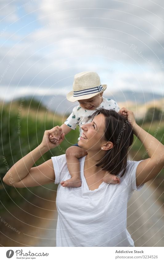 Adorable kid in hat on shoulders and looking at mother baby road maternity motherhood summer care casual laughing comfortable moment family child field parent