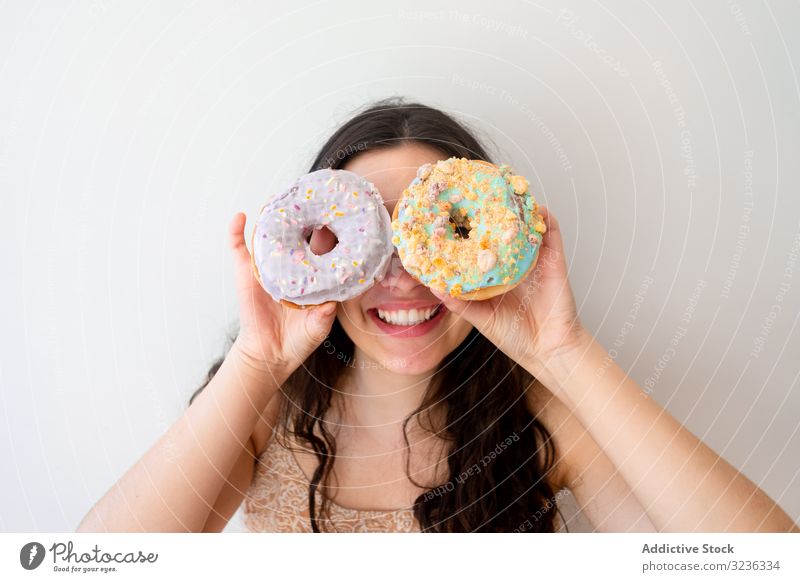 Playful woman making glasses of donuts while standing by white wall playful having fun carefree pastry glazed sprinkles relaxed funny young adult food enjoy