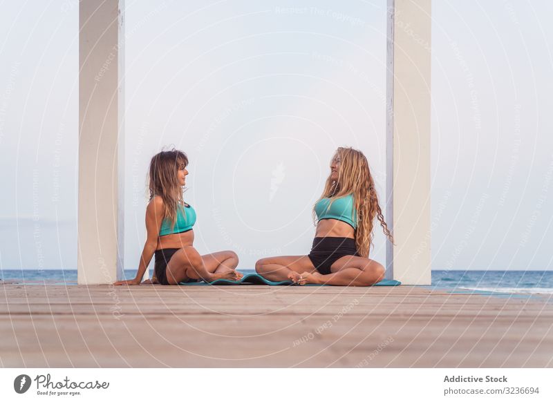 Smiling women sitting and stretching in butterfly posture at seaside acroyoga stretching butterfly exercise beach together partnership balance acrobatic fitness
