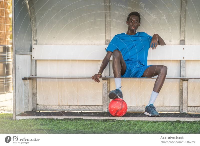 Black football player resting on bench teenager field sit break training sportswear ethnic male adolescent soccer sunny daytime black african american