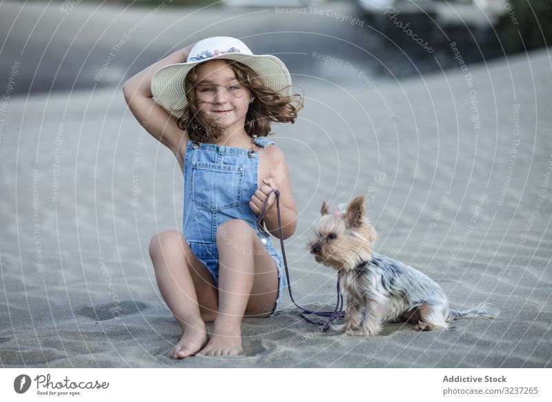 Cute girl holding hat and sitting on beach with dog pet sand summer sea animal friendship companion vacation fun shore nature windy together travel cute terrier