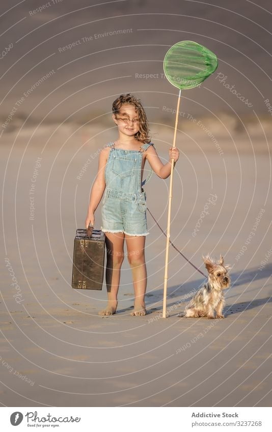 Curious kid with suitcase and butterfly net standing on sand with dog girl summer sea animal friendship pet companion vacation fun shore beach nature windy
