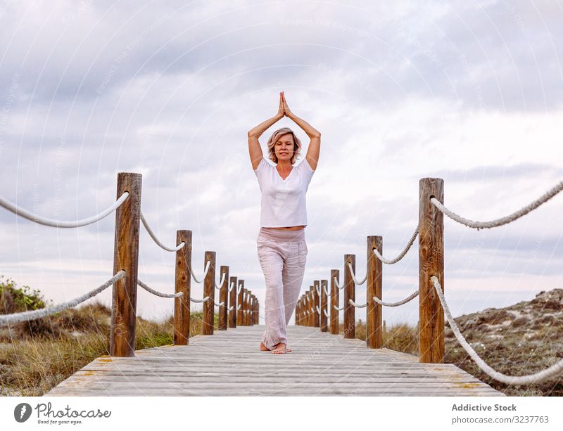 Cheerful lady exercising on timber path woman breath gesture exercise clasped hands tai chi meditation nature sky cloudy training barefoot female adult healthy