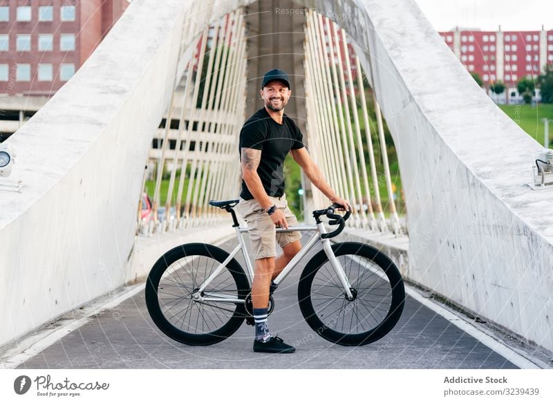 Cheerful man with bicycle standing on footbridge bike city ride modern active sportive summer male adult happy cheerful smile beard cap cyclist recreation