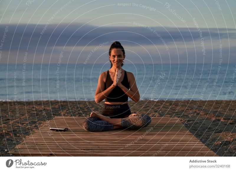 Adult woman meditating near sea in evening yoga beach asana activity fit clasped hands crossed legs sky cloudy female positive smile slim sportswear training