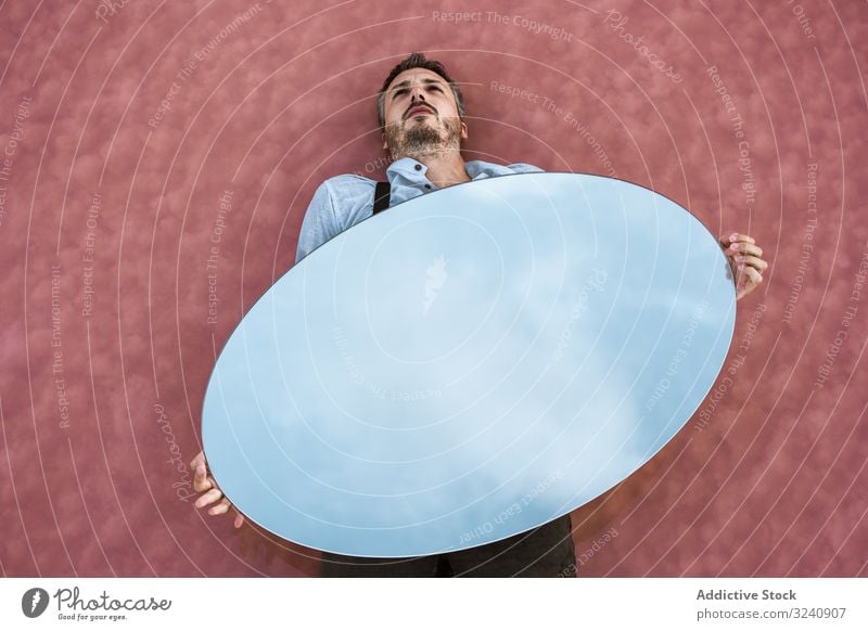 Man lying down holding mirror with reflection man stylish pensive white shirt suspenders blue sky carrying handsome oval surreal concept modern young adult art