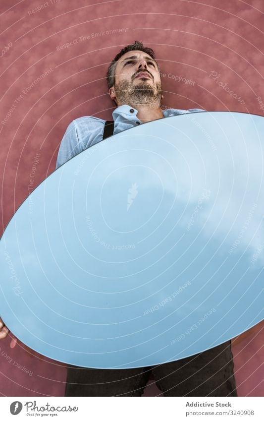 Man lying down holding mirror with reflection man stylish pensive white shirt suspenders blue sky carrying handsome oval surreal concept modern young adult art