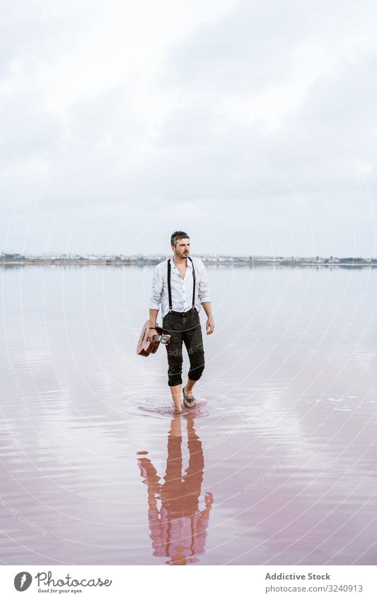 Musician holding guitar at seaside musician inspired barefoot stand play water cloudy man shore white shirt suspenders horizon reflection young guitarist