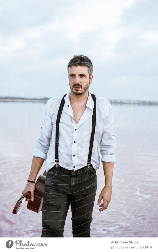 Musician holding guitar at seaside musician inspired barefoot stand play water cloudy man shore white shirt suspenders horizon reflection young guitarist