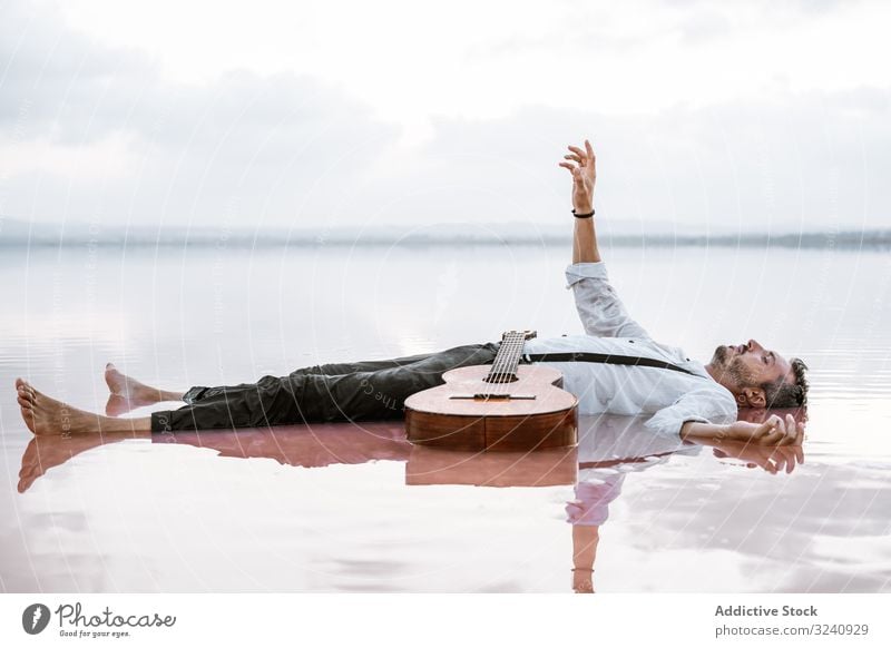 Man resting in water with guitar at seaside man classy lying on back floating sandbank white shirt suspenders dreamy peaceful calm neutral young adult bristle