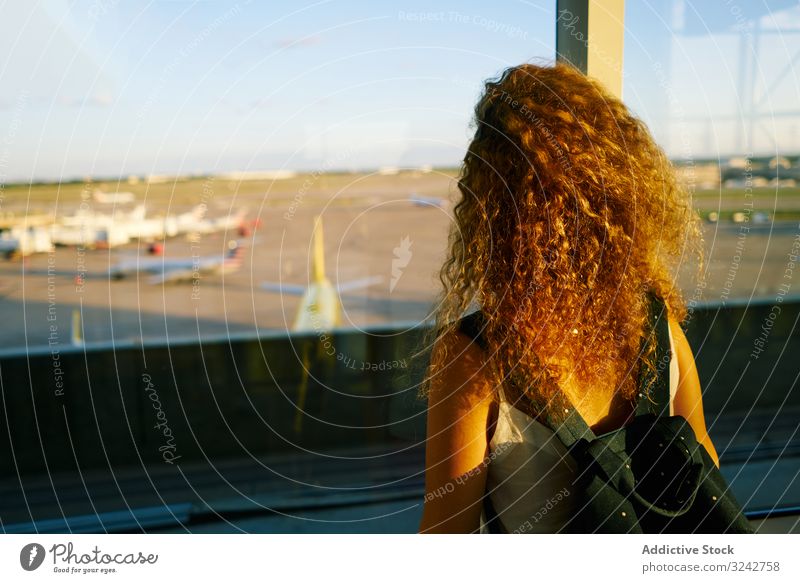 Young woman thoughtfully looking at airplanes through window in sunlight dream airport hall inspiration freedom anticipation observe travel flight backpack