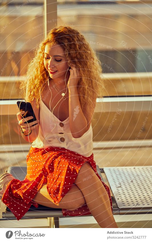 Smiling woman sitting on metal bench with earphones and smartphone music airport listen waiting area female casual style texas using selfie relax modern rest
