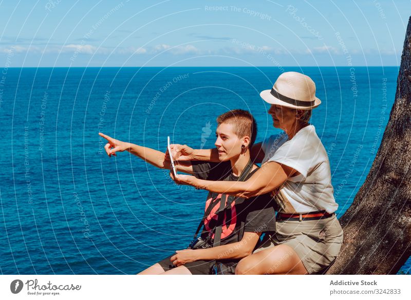 Stylish woman with tablet in hands and elderly lady women mature sea rocky finger pointing using water blue sky vacation summer ocean happy tropical beach
