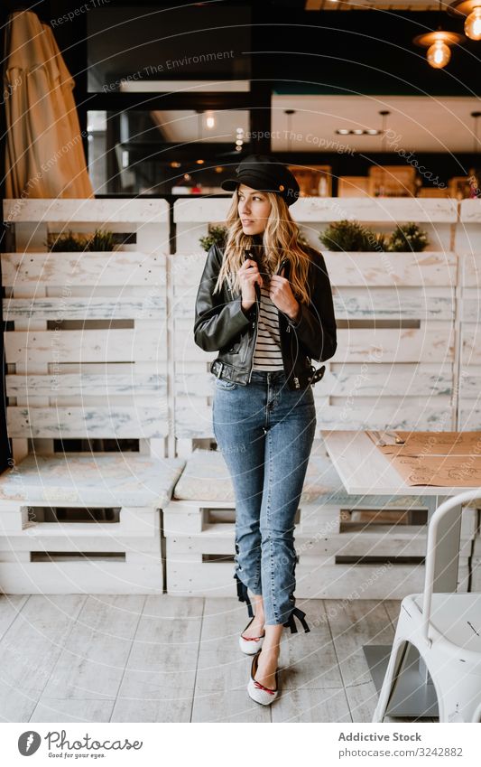 Casual stylish woman standing at table in cafe waiting design wooden trendy blond creative clothing leather jacket casual denim bright curly model female vogue