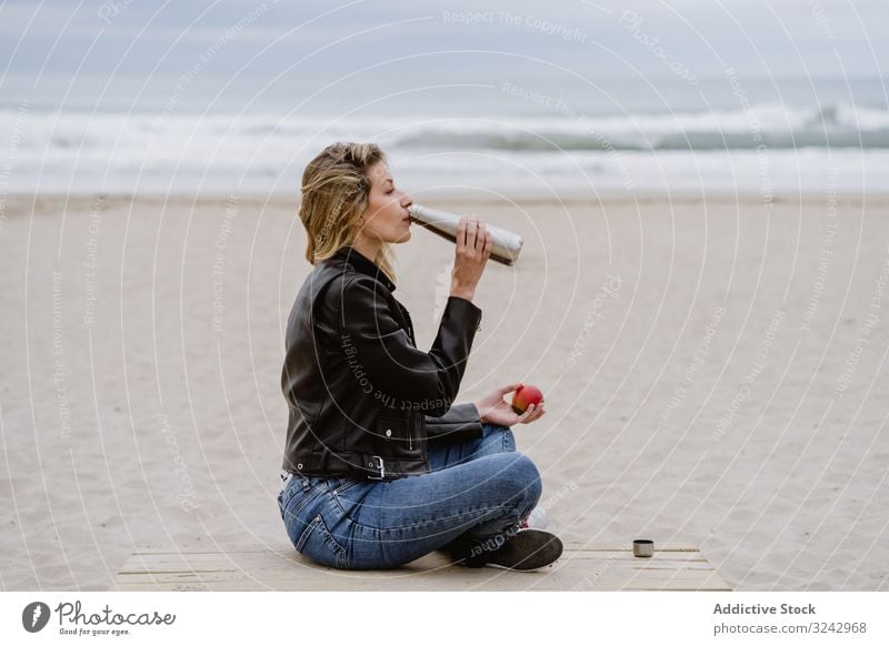 Content woman drinking and eating apple sitting with crossed legs on beach snack healthy lifestyle food fruit pier bottle warm steel reusable eco friendly
