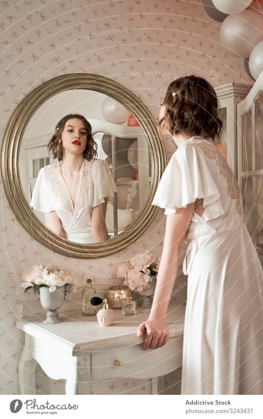 Attractive serious young woman standing and looking in big round mirror in boudoir reflection sensual allure wedding dress red lips beauty silk makeup chic